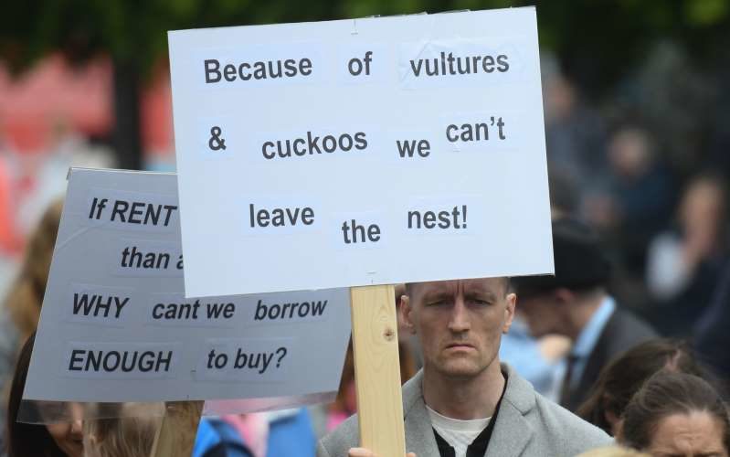 man holds a sign which reads "Because of vultures & cuckoos we can't leave the nest!"